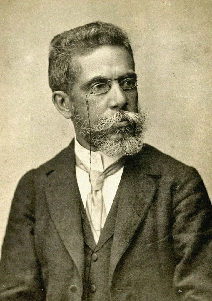 Fotografia do escritor Machado de Assis. Ele está sentado em pose, olhando acima do ombro, utilizando óculos, terno e gravata, além de uma barba avantajada e bigode característico.